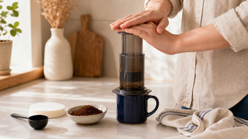 Hands press an AeroPress over a navy mug beside filters and measured coffee grounds.