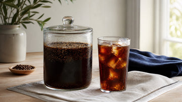 A glass jar of coarse coffee grounds steeps beside a finished glass of cold brew over ice.