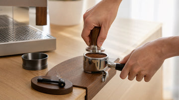 Hands tamp a level coffee puck in a portafilter beside a WDT distribution tool.