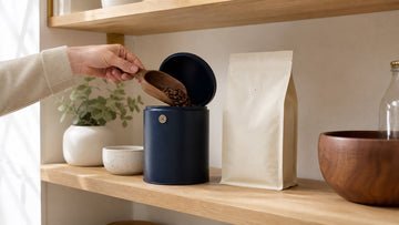 Whole coffee beans are placed into an airtight navy canister beside a valve coffee bag.