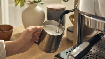A stainless pitcher is held beneath a steam wand while glossy microfoam swirls inside.