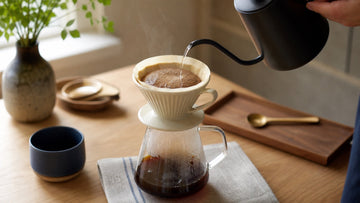 A gooseneck kettle pours water over freshly ground coffee in a ceramic pour-over dripper above a glass carafe.