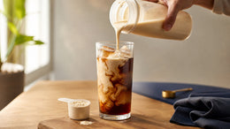 A creamy protein shake is poured into cold brew over ice beside a scoop of protein powder.